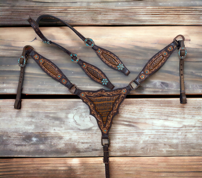 A leather headstall and breast collar set with intricate filigree carving and light brown alligator leather accents, laid out on a wooden surface.
