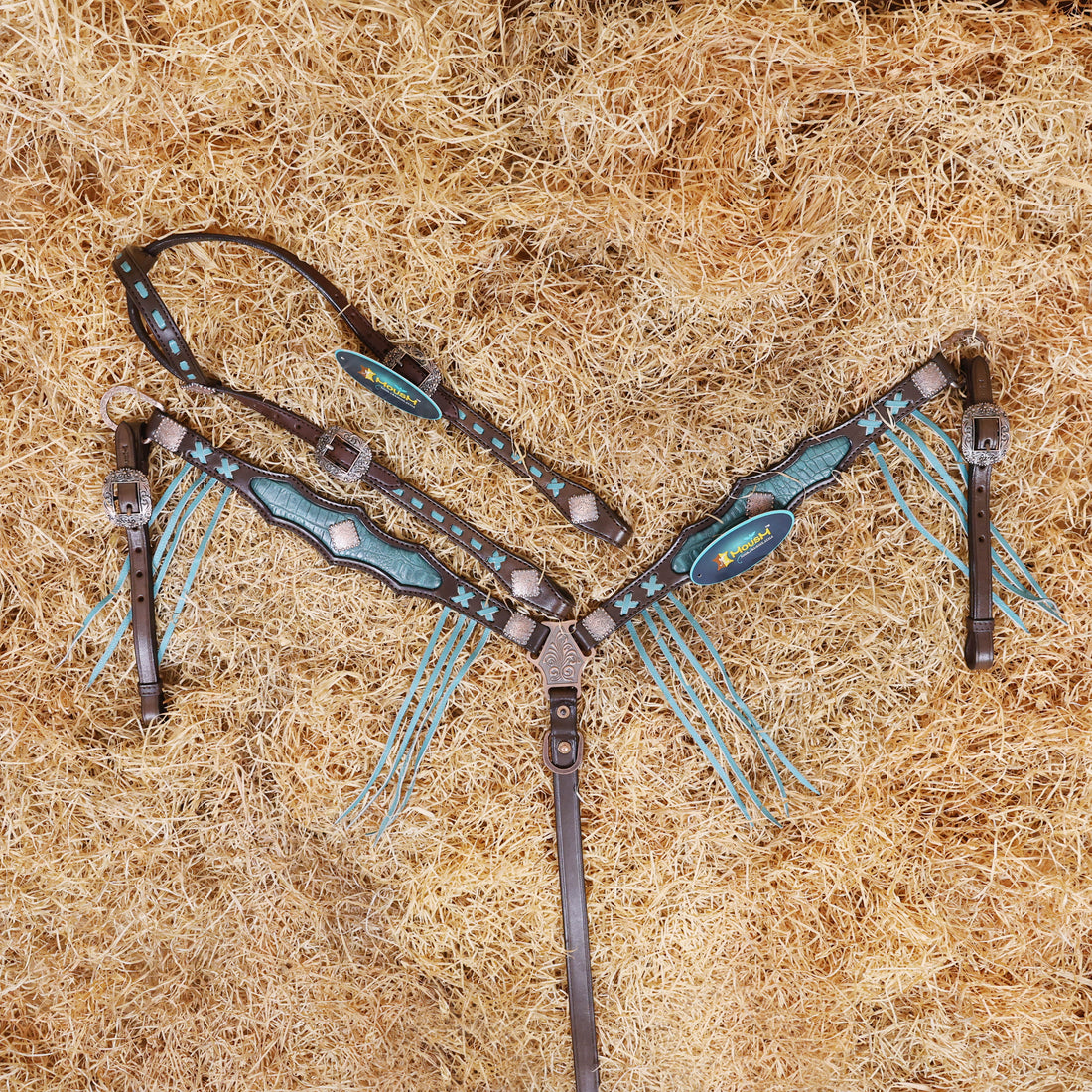 A Western horse bridle and breast collar set with turquoise croc print cowhide leather inlay and fringe, laid out on a hay surface.