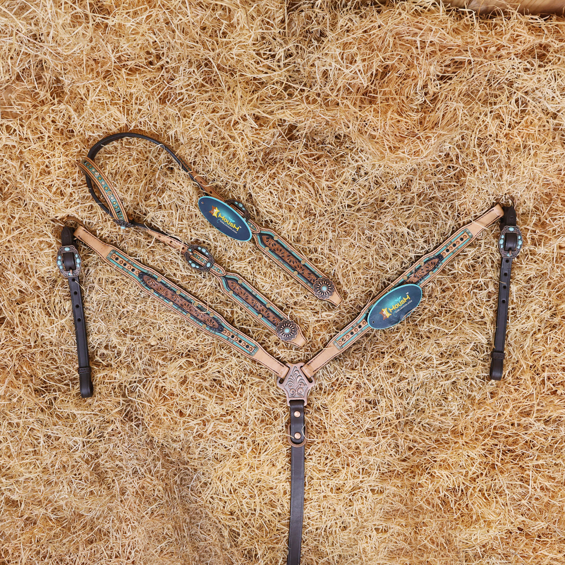 A tooled leather Western headstall and breast collar set with floral design, featuring topaz and black rivets, and antique copper hardware, laid out on a bed of straw.