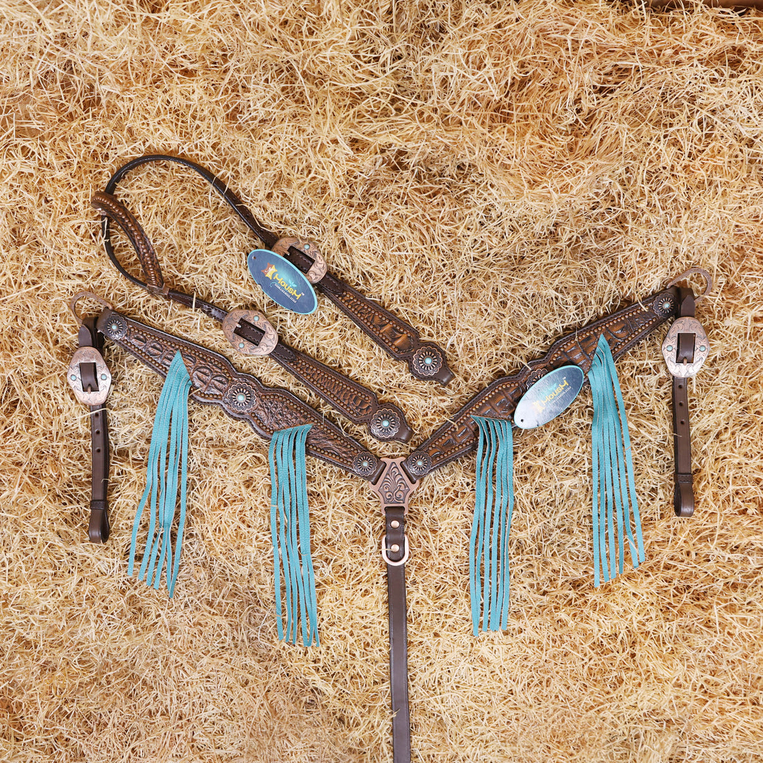A set of Argentina cow leather Western horse tack including a bridle and breast collar with 3D croc print, turquoise fringe, copper studs, and antique copper hardware, displayed on a bed of straw.