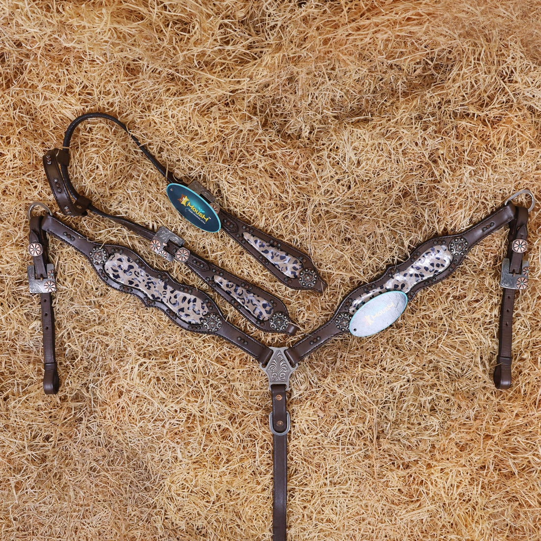 A set of Argentina cow leather horse tack, featuring a headstall and breast collar with leopard printed leather decorations and antique copper hardware, displayed on a bed of straw.