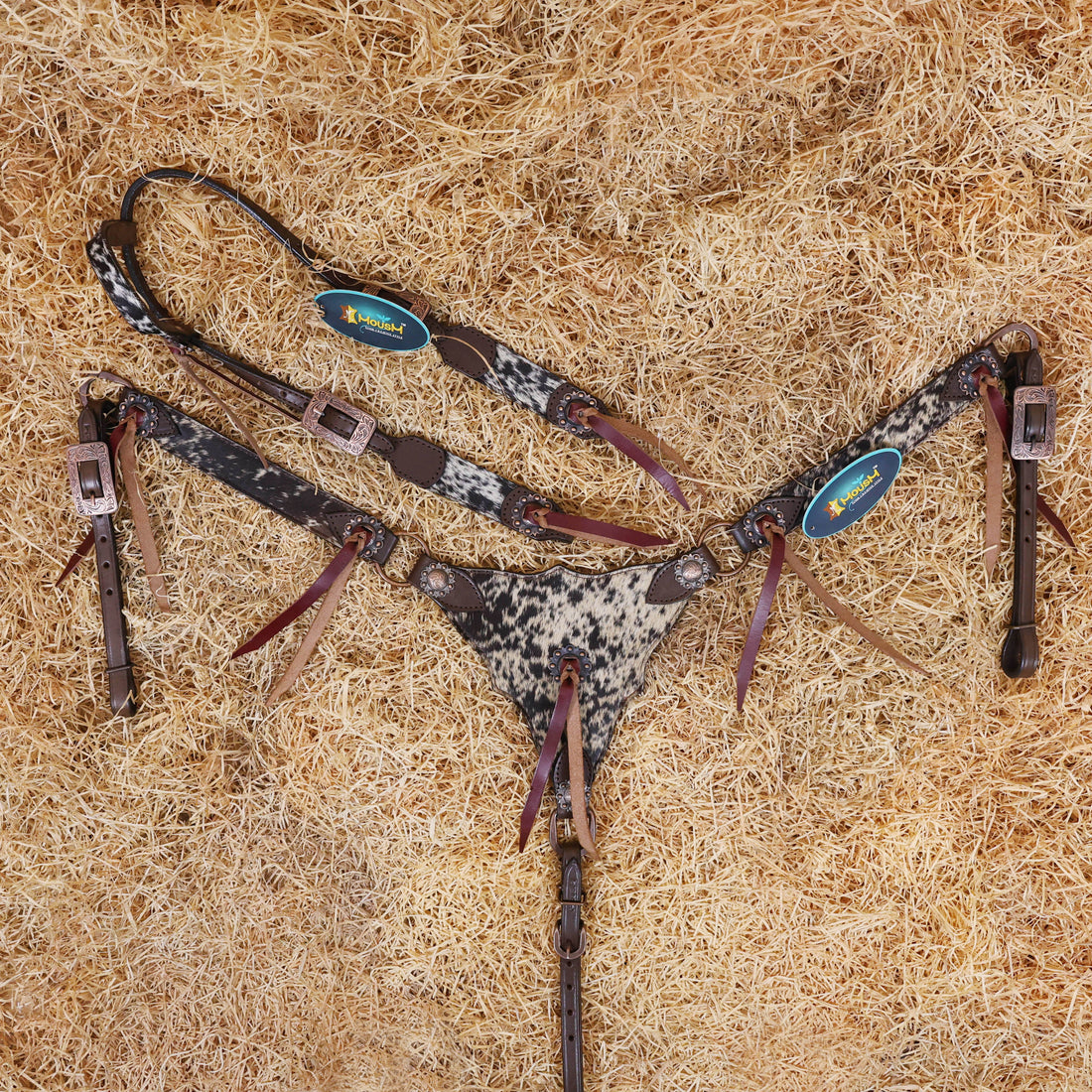 A set of Argentina cow leather Western horse tack including a bridle and breast collar with natural salt paper, cowhide, and string concho details, laid out on a bed of straw.